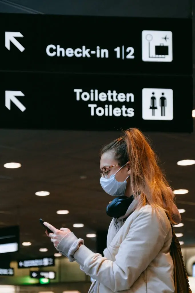 Young woman wearing a medical mask to protect against coronavirus, waiting at the airport and typing on mobile phone 